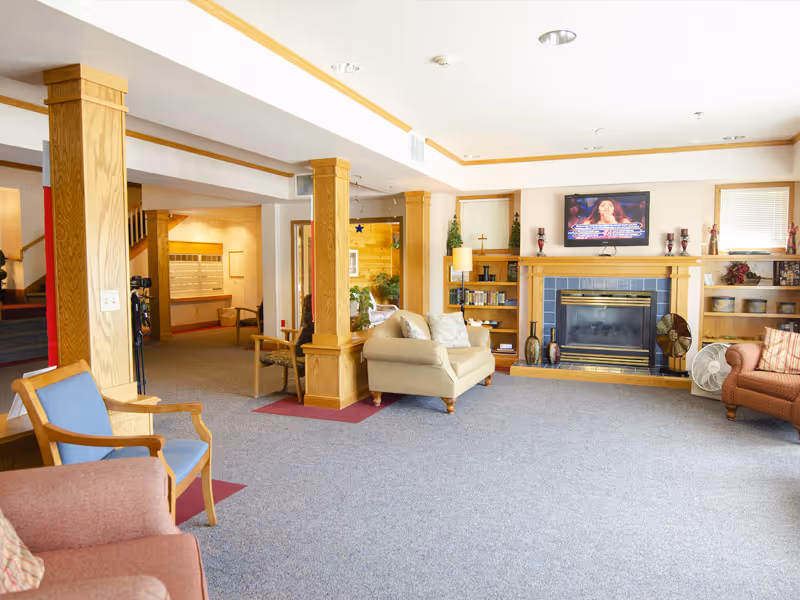 A spacious living room area in a senior living facility with beige and brown furniture including armchairs and a loveseat. The room features wooden columns, built-in shelves with books and decorative items, a fireplace with a TV mounted above it, and carpeted flooring. There are windows with blinds letting in natural light.