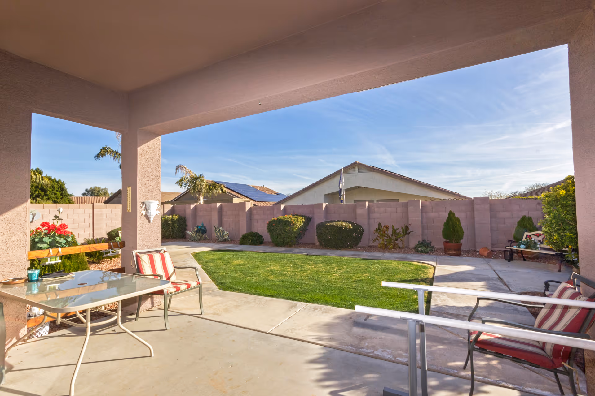 Covered patio area with a glass table and striped cushioned chairs overlooking a small, well-maintained grassy yard with bushes and a concrete walkway, surrounded by a brick privacy wall and neighboring houses under a clear blue sky.