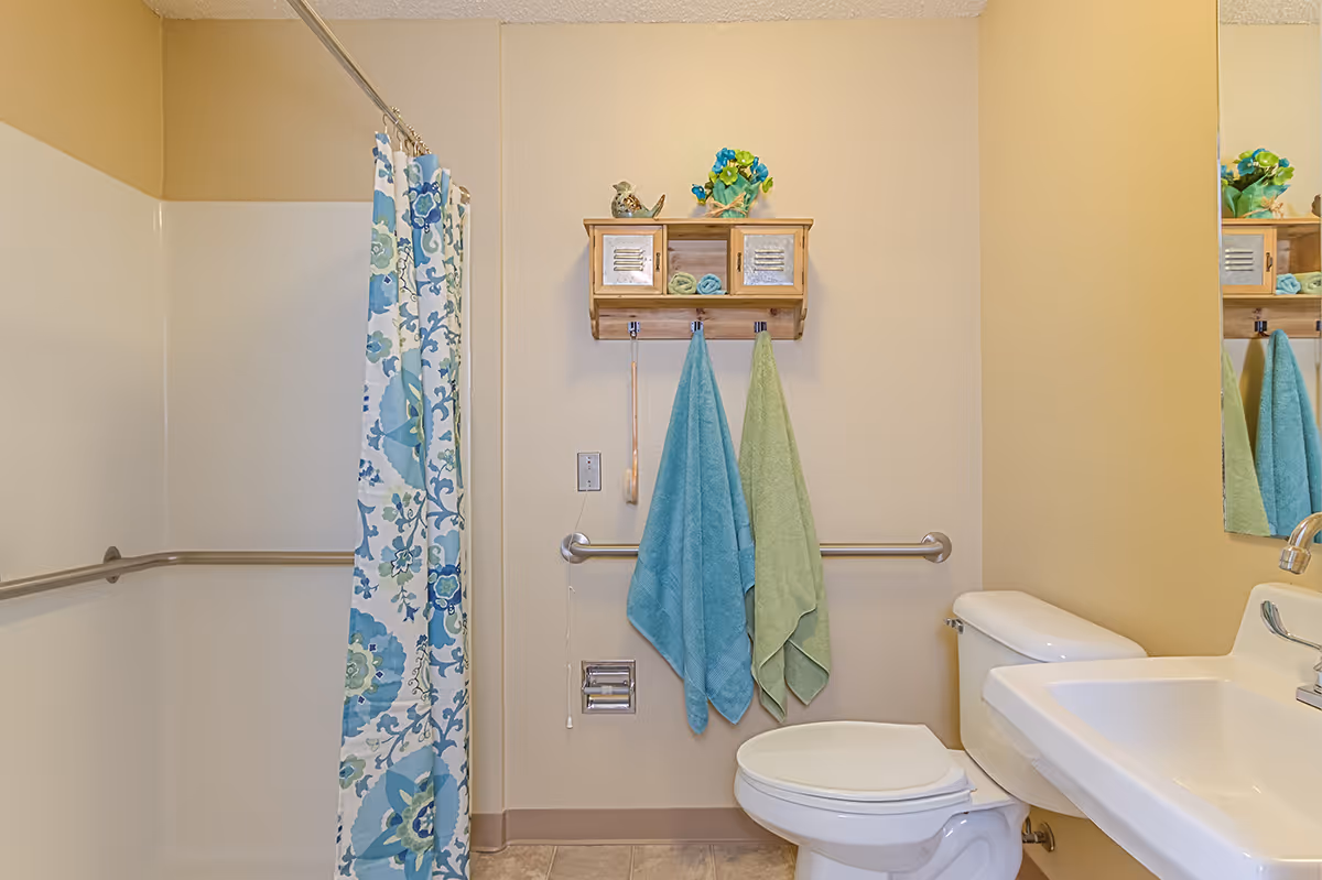 A clean bathroom with a white toilet and sink, a shower with a blue and green floral patterned curtain, two towels hanging on hooks on the wall, and a small wooden cabinet with decorative items and rolled towels above the towels.