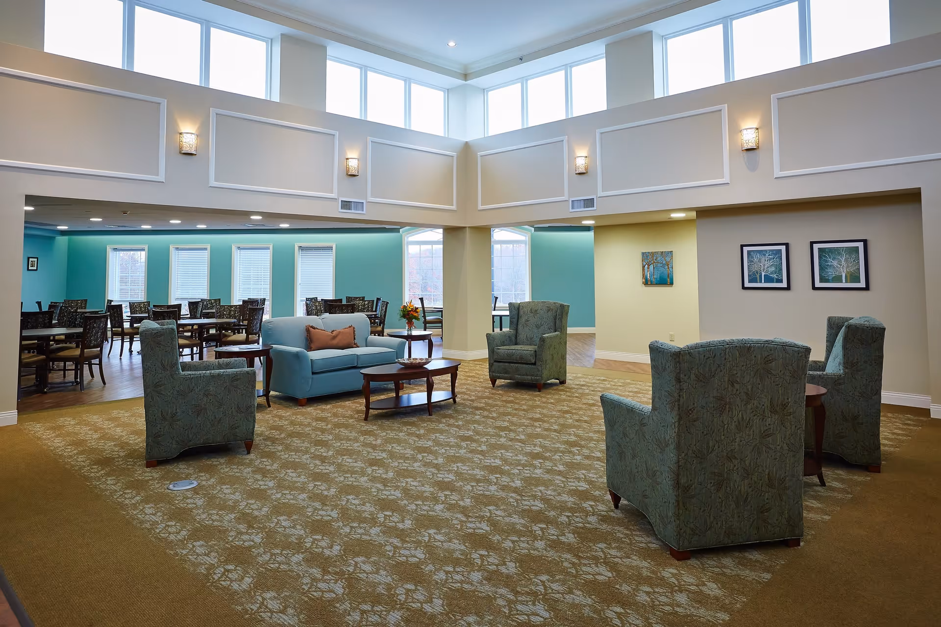 A spacious senior living common area with patterned carpet, four green upholstered armchairs arranged around a small wooden table, and a light blue sofa with a brown cushion. The room has high ceilings with large windows near the ceiling allowing natural light. In the background, there is a dining area with multiple tables and chairs, teal-colored walls, and several windows with blinds. The walls in the common area are beige with framed artwork and wall sconces providing additional lighting.