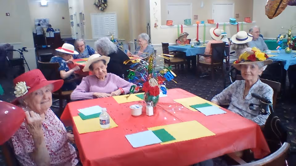 A group of elderly women wearing colorful hats sitting around tables covered with red and blue tablecloths in a communal dining area. The tables have festive centerpieces and place settings with napkins and cups. The room has a warm, inviting atmosphere with other seniors seated in the background.