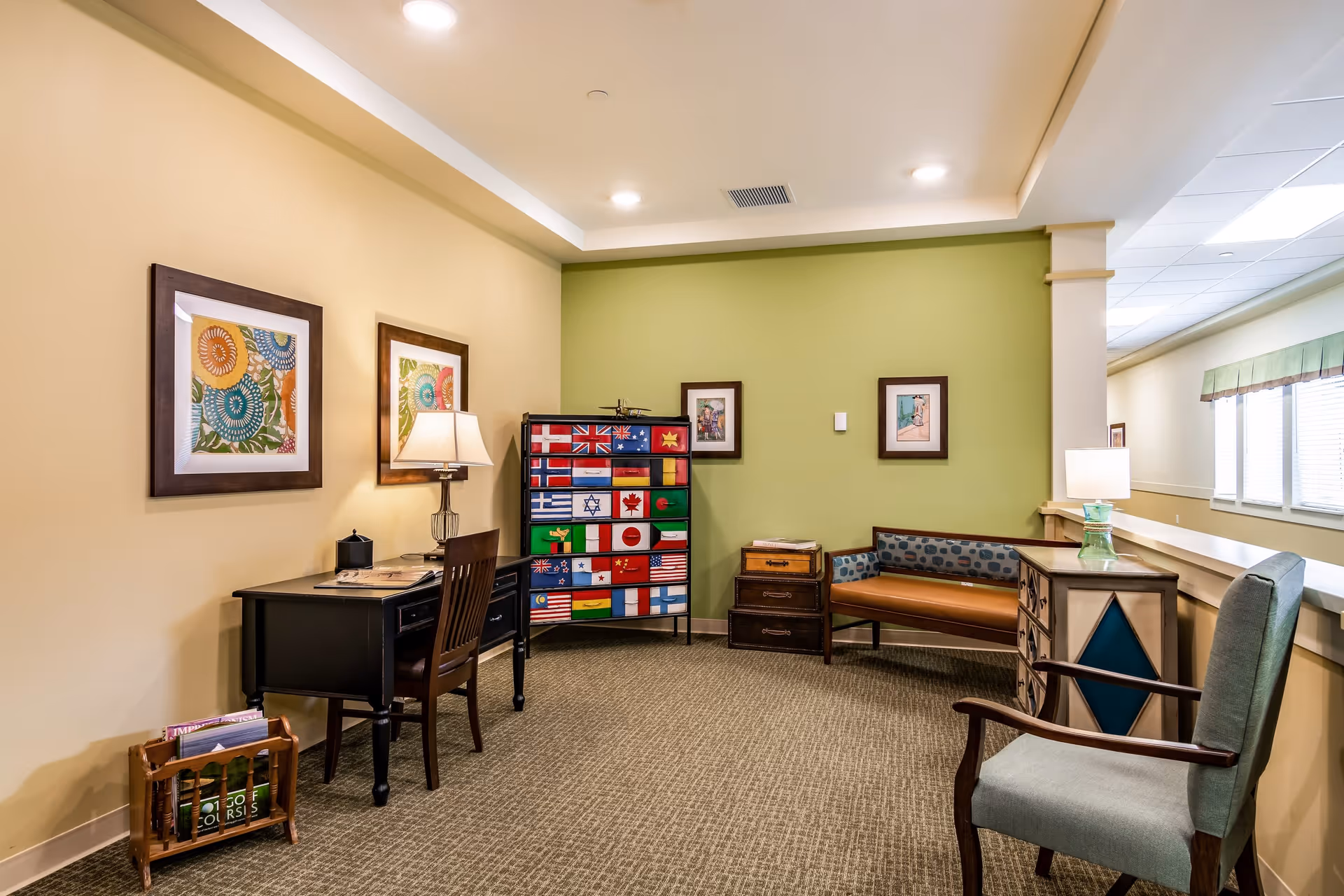 A cozy interior room with beige and green walls, featuring a black desk with a chair and a lamp, colorful framed artwork on the walls, a chest of drawers decorated with various international flags, a small wooden bench with patterned cushions, and an upholstered armchair. The room is well-lit with ceiling lights and natural light from windows on the right side.