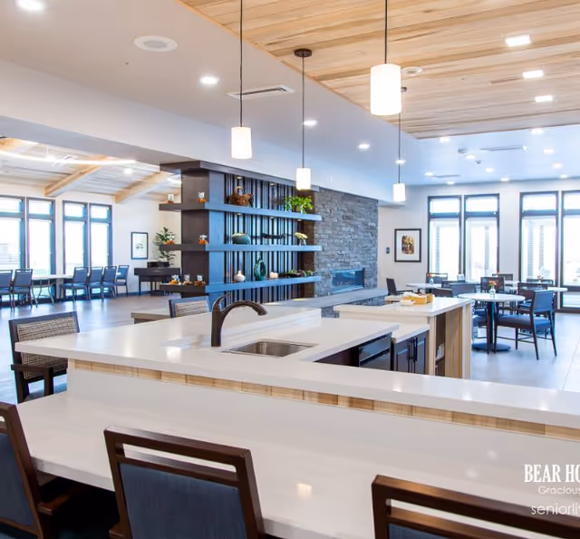 A spacious, modern common area in a senior living facility featuring a large kitchen island with a sink and faucet, surrounded by chairs. The room has pendant lights hanging from a wooden ceiling, a stone accent wall with a fireplace, and multiple tables and chairs near large windows allowing natural light to fill the space.