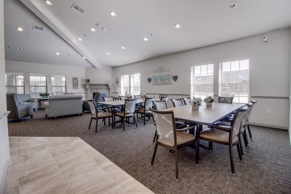 A bright and spacious common area in an assisted living facility featuring two rectangular tables with chairs arranged around them, small potted plants on the tables, and a seating area with sofas and armchairs near a fireplace. Large windows allow natural light to fill the room, and decorative wall art is visible.