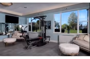 A fitness room with exercise equipment including a multi-function weight machine, a wall-mounted TV, large windows showing a green outdoor area, and comfortable seating with a beige sofa and round ottoman.