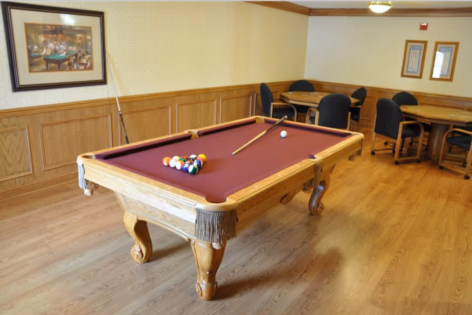 A recreational room with a wooden pool table featuring a maroon felt surface, pool balls racked and a cue stick resting on the table. The room has wooden flooring, wood-paneled lower walls, and wallpapered upper walls. In the background, there are round tables with black cushioned chairs and framed pictures on the walls.