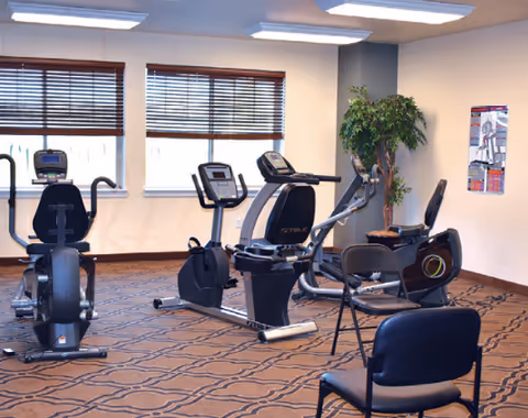 A fitness room with exercise equipment including stationary bikes and an elliptical machine. The room has patterned carpet, two windows with wooden blinds, a potted plant in the corner, and a chair in the foreground. A poster is visible on the wall.