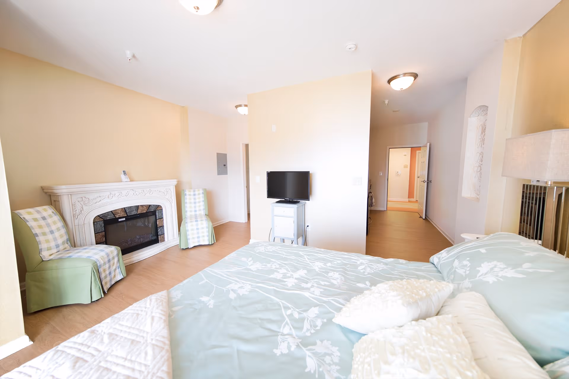 A bright bedroom with a bed covered in light blue floral bedding and white pillows. Two green chairs with plaid covers are positioned near a decorative white fireplace. A small TV sits on a white stand against a beige wall. The room has light wood flooring and a hallway leading to an open door in the background.