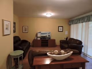 A cozy living room with beige walls, a ceiling light, and a large window covered by a curtain with a valance. The room contains two dark brown leather sofas, a wooden coffee table with a decorative bowl filled with natural elements, a small round side table, and a wooden entertainment center with a stereo system and framed pictures on the wall.