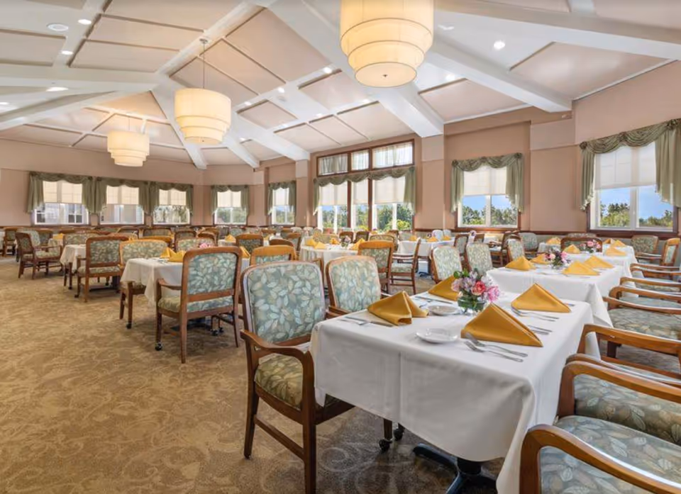 A spacious dining room with multiple tables covered in white tablecloths, each set with yellow folded napkins, plates, and silverware. The chairs have wooden frames with green patterned upholstery. Large windows with green curtains allow natural light to fill the room. The ceiling features exposed white beams and large round pendant lights.