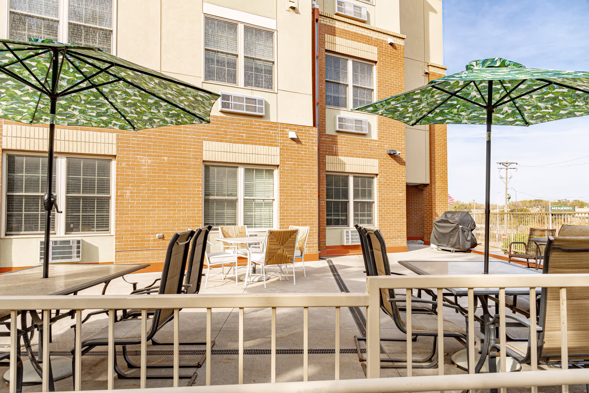 Outdoor patio area with several tables and chairs under large green umbrellas with leaf patterns. The patio is adjacent to a brick and beige building with multiple windows and air conditioning units. A covered grill is visible in the background along with a fence and some distant trees under a clear sky.