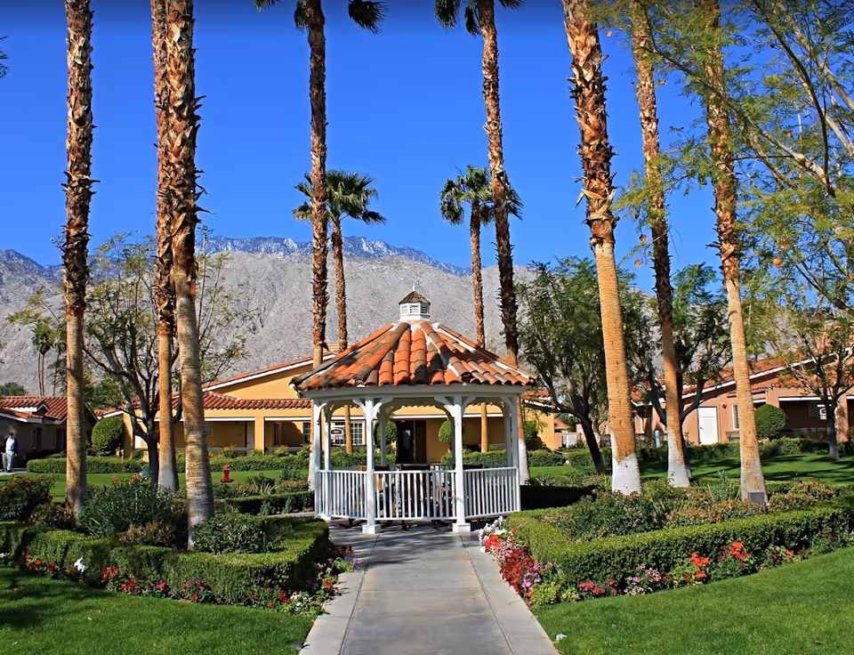 A landscaped outdoor area at Cottages at Palm Springs featuring a white gazebo with a red-tiled roof, surrounded by neatly trimmed hedges, colorful flowers, tall palm trees, and green grass. In the background, there are single-story buildings with red-tiled roofs and a mountain range under a clear blue sky.