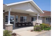 Front exterior view of a single-story building with a covered porch supported by white columns. The porch has several black chairs lined up against the wall. The building features brick and siding exterior with multiple windows and a well-maintained garden area in front.