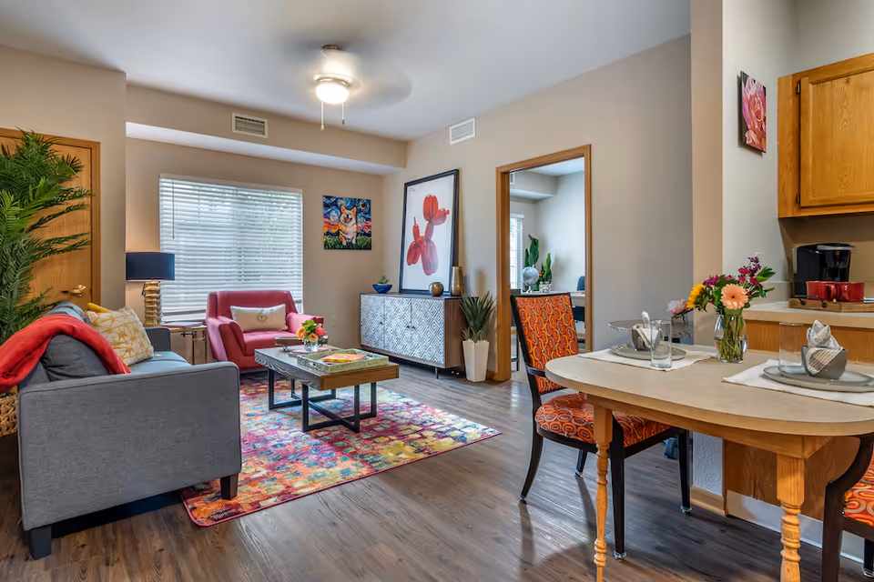 A cozy senior living facility interior featuring a living room and dining area. The living room has a gray sofa with colorful pillows and a red throw, a pink armchair, a wooden coffee table on a vibrant multicolored rug, and a sideboard with decorative items and artwork on the walls. The dining area has a small wooden table set for two with orange patterned chairs and a vase of flowers. The kitchen area with wooden cabinets and a coffee maker is partially visible.