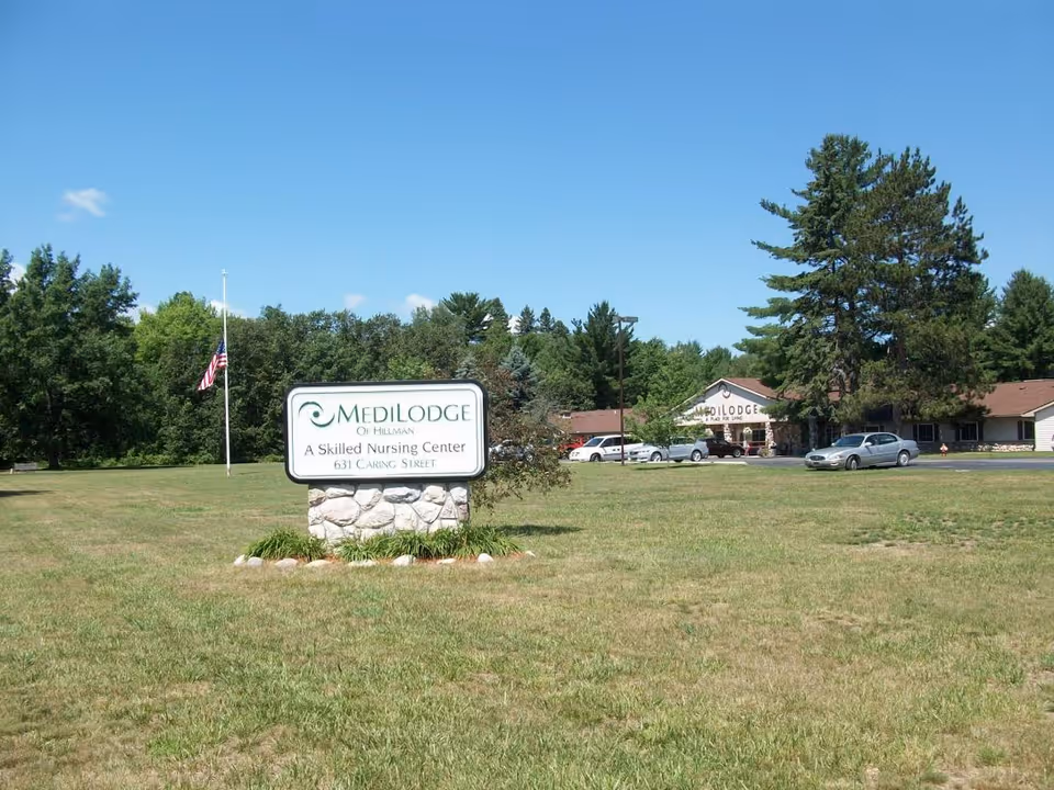 A Medilodge of Hillman sign on a stone base in front of a grassy lawn and the nursing center building under a clear blue sky.