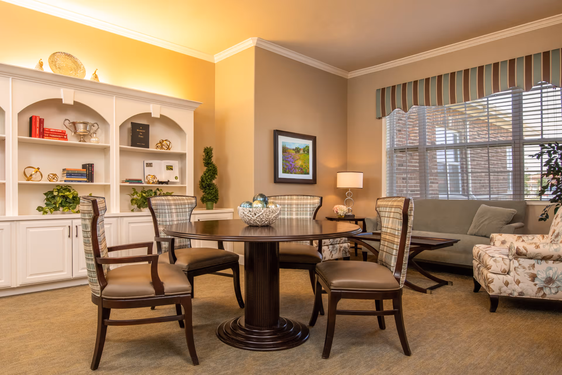 A cozy interior room with a round wooden table surrounded by four chairs with plaid upholstery. Behind the table is a white built-in bookshelf with decorative items and books. To the right, there is a sitting area with a green sofa, a floral armchair, a wooden coffee table, a side table with a lamp, and a large window with striped valance curtains letting in natural light.
