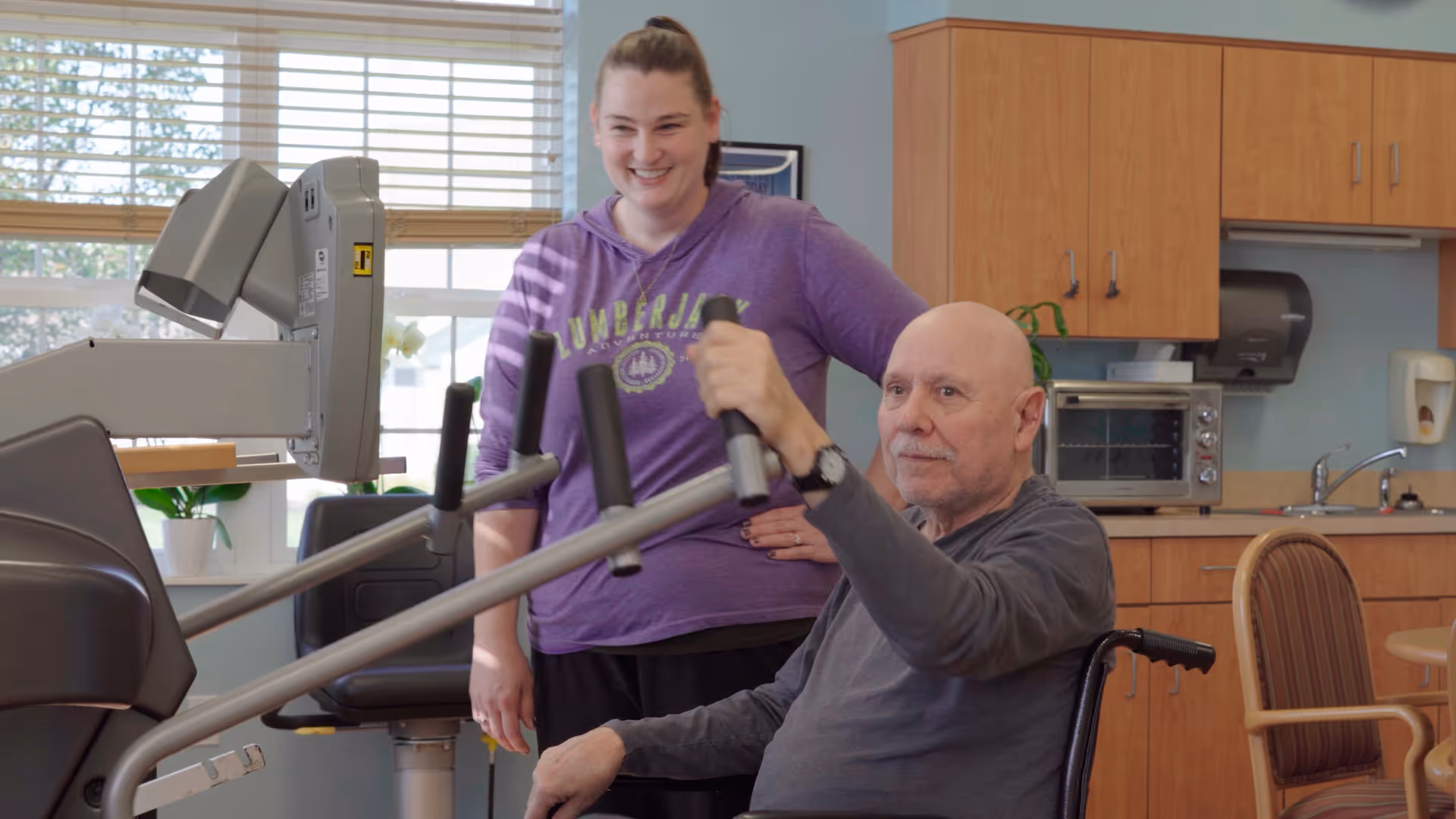 An elderly man in a wheelchair is using an exercise machine in a room that looks like a common area or therapy room. A woman in a purple sweatshirt stands behind him, smiling and providing support. The room has wooden cabinets, a countertop with a toaster oven, a sink, and a window with blinds letting in natural light.