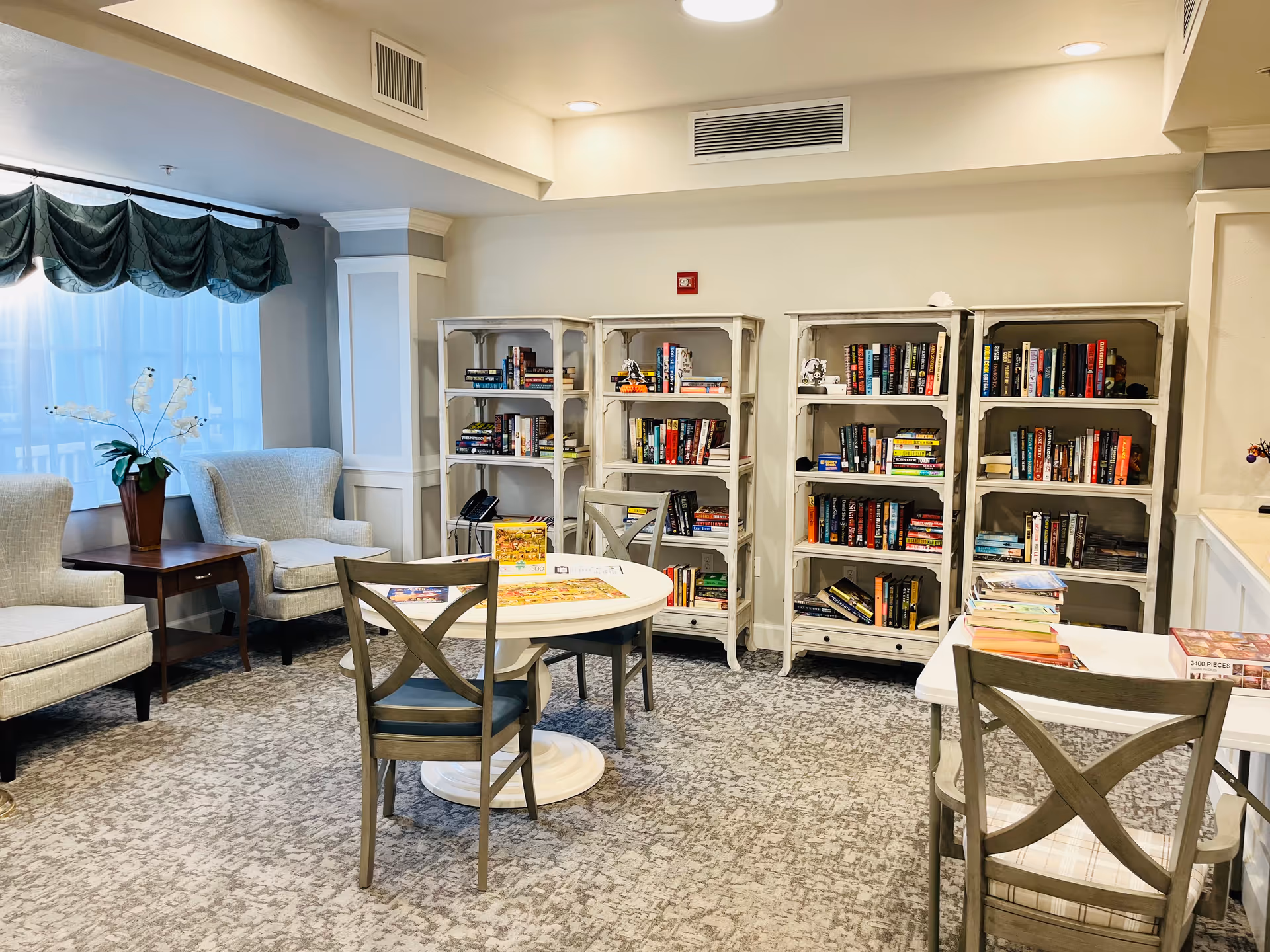A cozy common room with bookshelves, round tables and chairs, and upholstered armchairs by a curtained window.