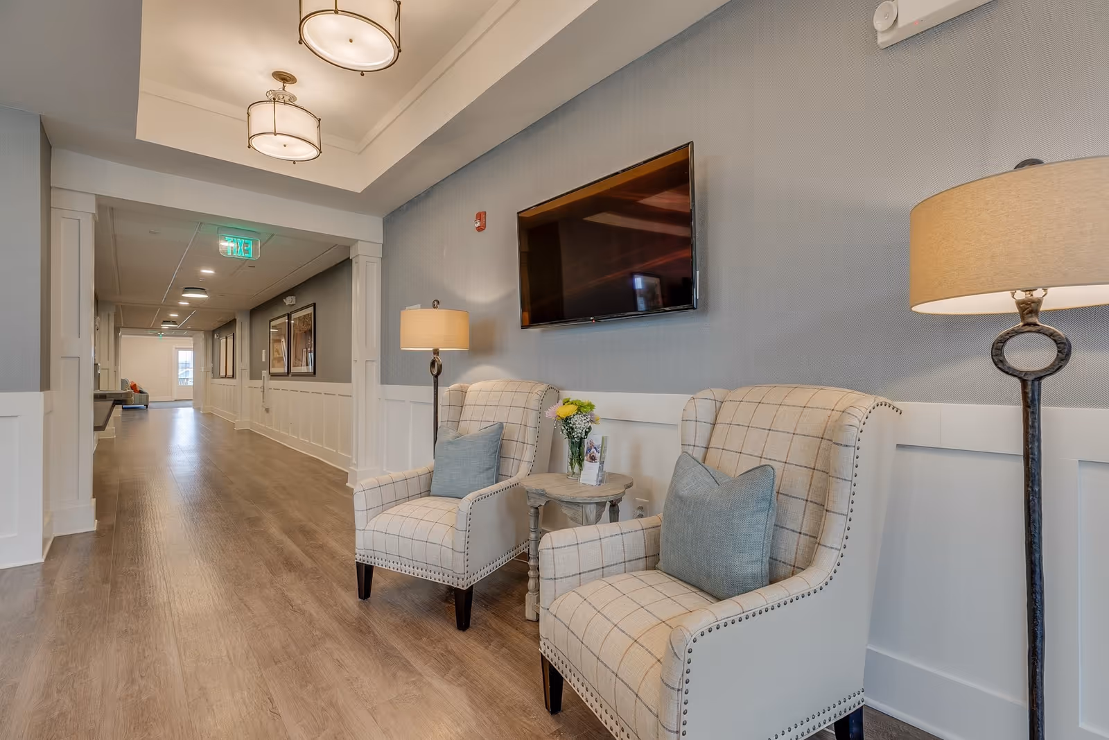 A well-lit hallway in a senior living facility with two upholstered armchairs featuring plaid patterns and blue cushions, a small round wooden table with a vase of flowers between them, a floor lamp, and a wall-mounted flat-screen TV. The hallway has wood flooring, light gray walls with white wainscoting, ceiling lights, and framed artwork along the corridor.