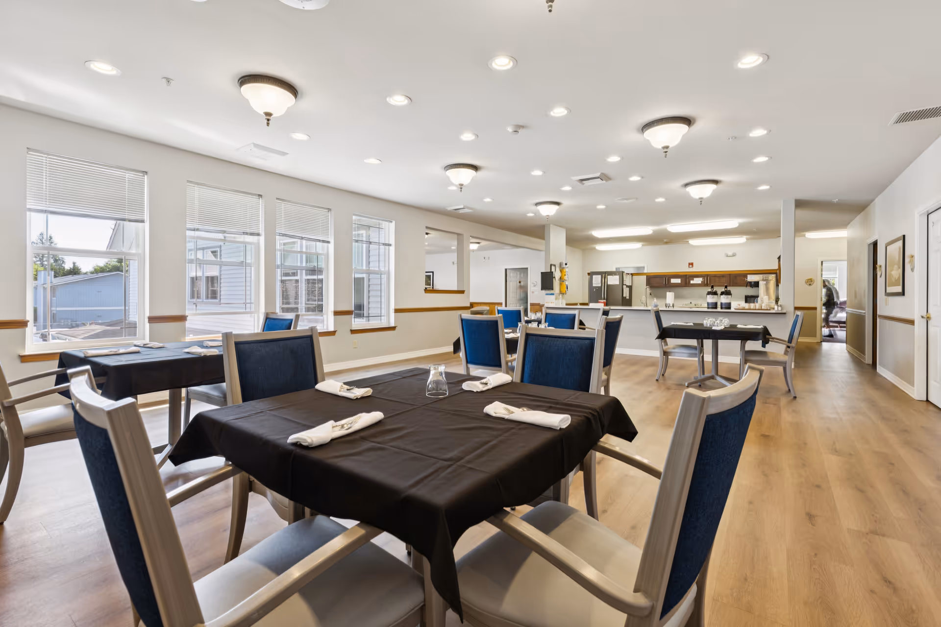 A bright dining room with several tables covered in black tablecloths, each set with napkins and glassware. The room has large windows letting in natural light, blue cushioned chairs, and a kitchen area in the background with coffee dispensers and cabinets.