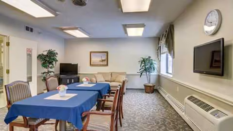 Communal dining room with blue tablecloths, chairs, sofa, plants, and wall-mounted TV.