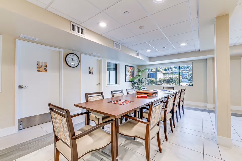 A bright and clean common room with a long wooden table surrounded by chairs. On the table, there are board games including checkers and a stack of other games. The room has large windows letting in natural light, a clock on the wall, and a potted plant in the corner. The walls are light-colored, and the floor is tiled.