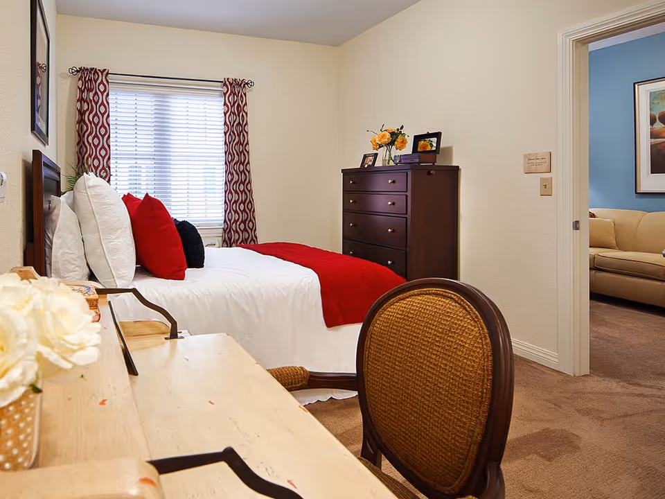 A cozy bedroom with a bed dressed in white linens and red and black pillows, a wooden dresser with framed photos and a vase of yellow flowers, a window with patterned red curtains, and a wooden desk with a chair in the foreground. An open doorway reveals a beige sofa in an adjacent room with blue walls.