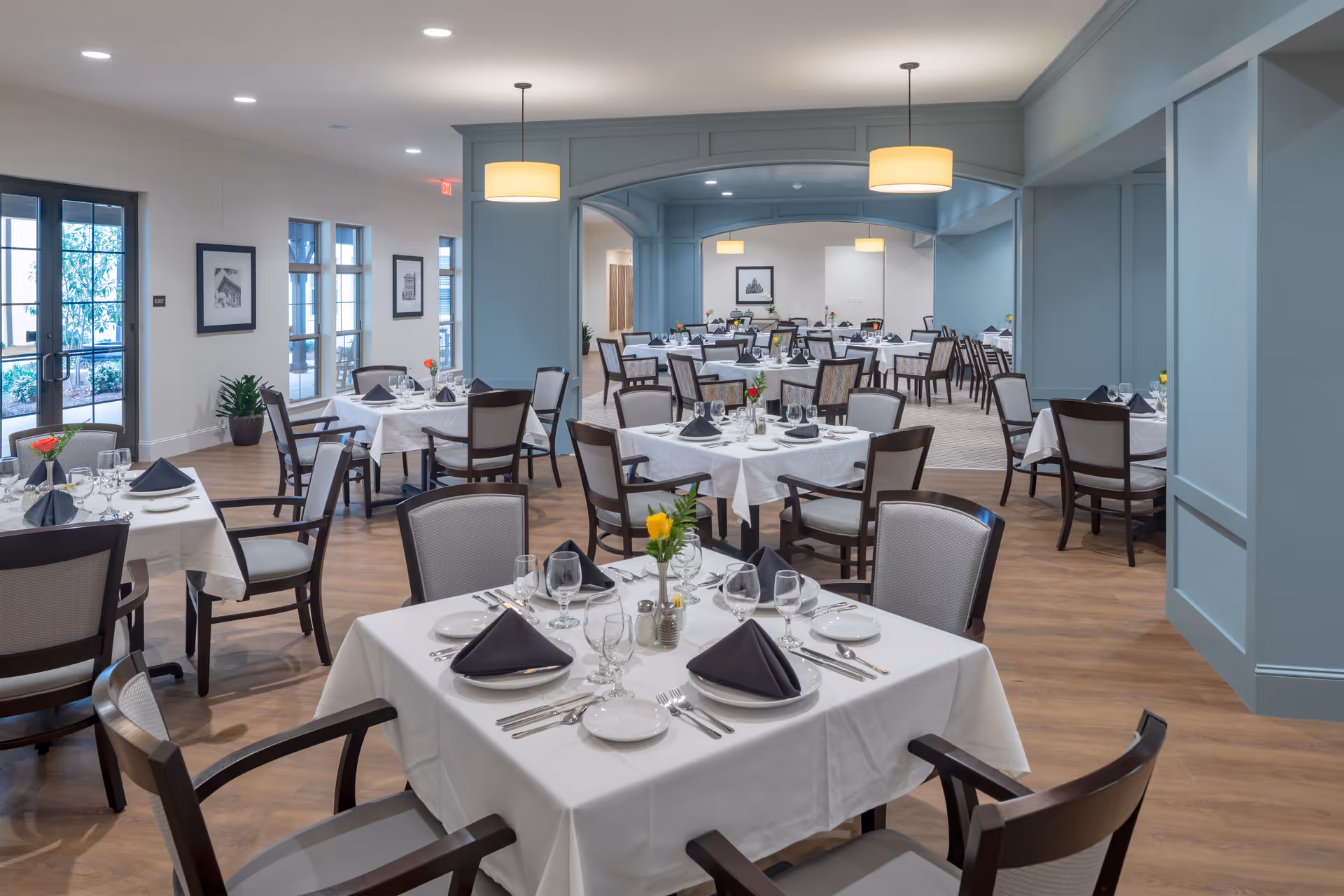 A spacious dining room with multiple tables covered in white tablecloths, each set with plates, glasses, silverware, and black folded napkins. The room features wooden flooring, light blue walls with decorative paneling, and pendant lights hanging from the ceiling. There are framed pictures on the walls and large windows letting in natural light.