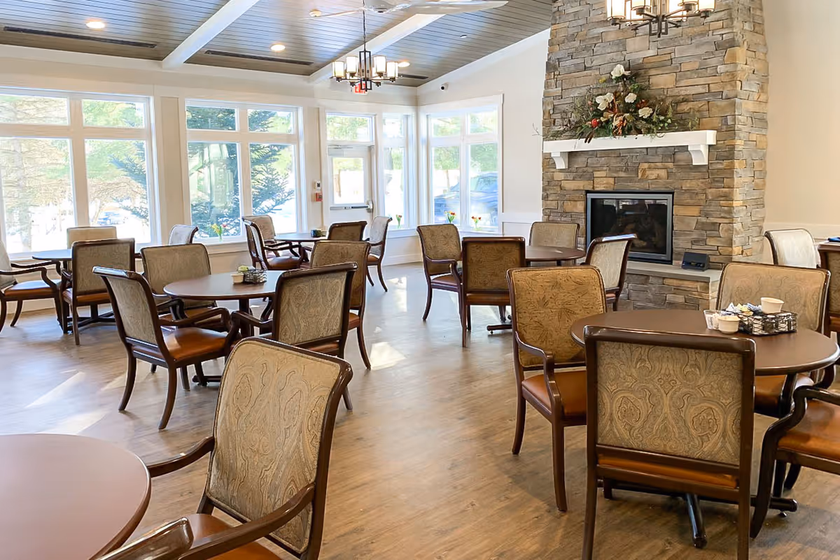 A bright dining room with multiple round tables and cushioned chairs arranged neatly. Large windows allow natural light to fill the space, and a stone fireplace with a floral arrangement on the mantel adds a cozy touch. The ceiling features wooden paneling and recessed lighting.