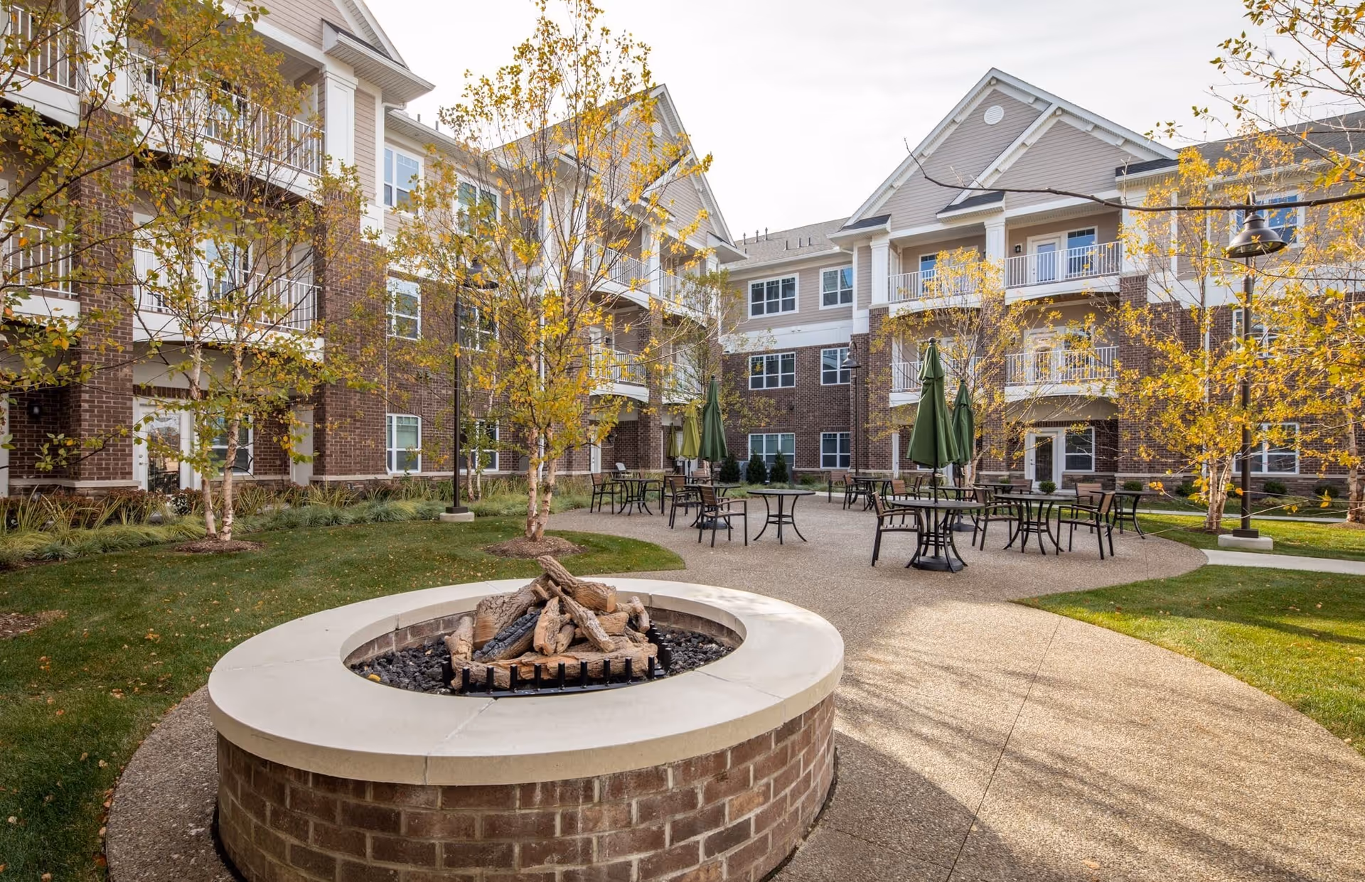 Courtyard of a senior living complex with a central fire pit, outdoor tables and umbrellas, and a brick multi-story building in the background.