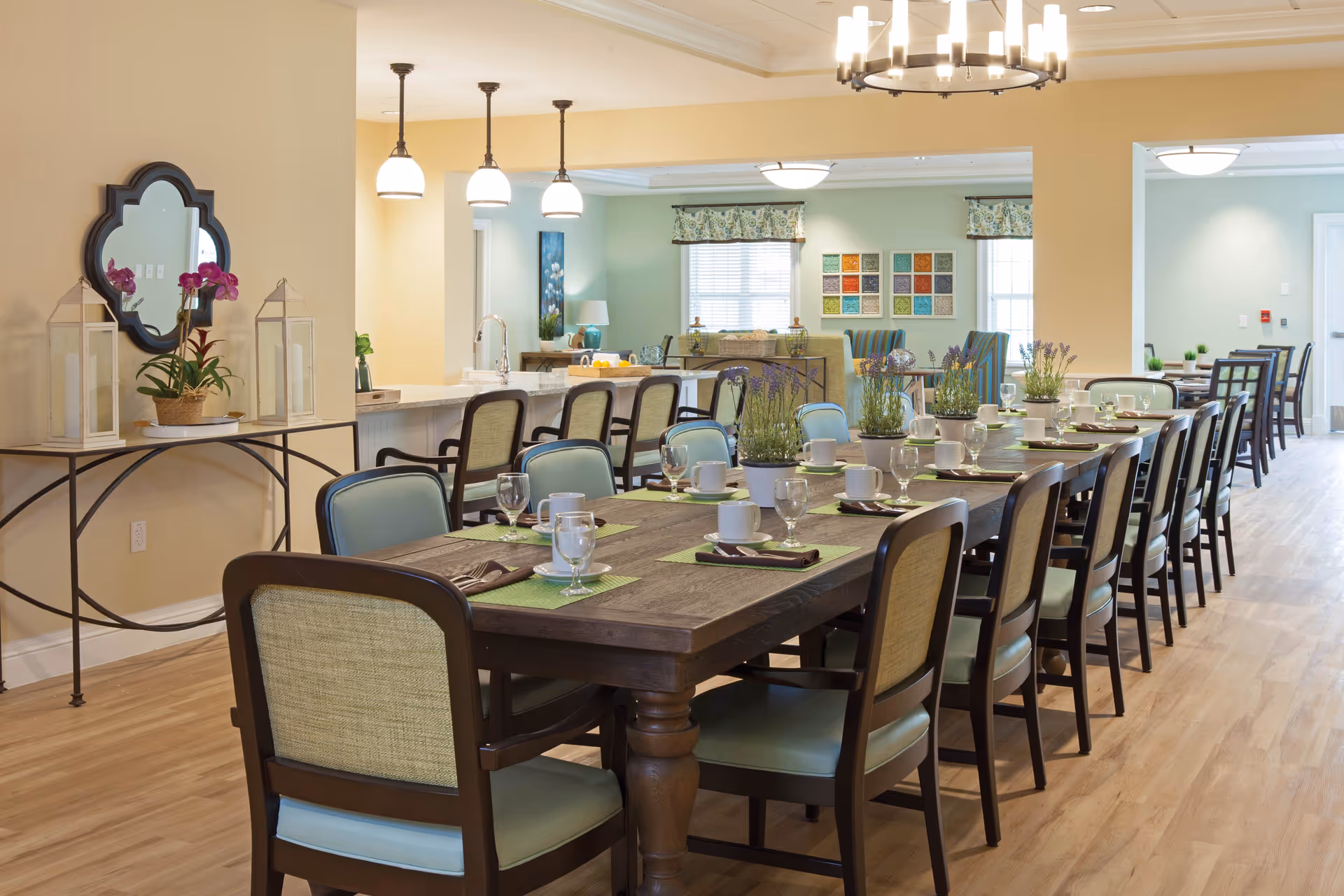 A long dining table set with cups, glasses, and napkins in a well-lit dining room. The room features wooden flooring, multiple chairs with light blue cushions, pendant lights above a counter with bar stools, and decorative plants and artwork on the walls.