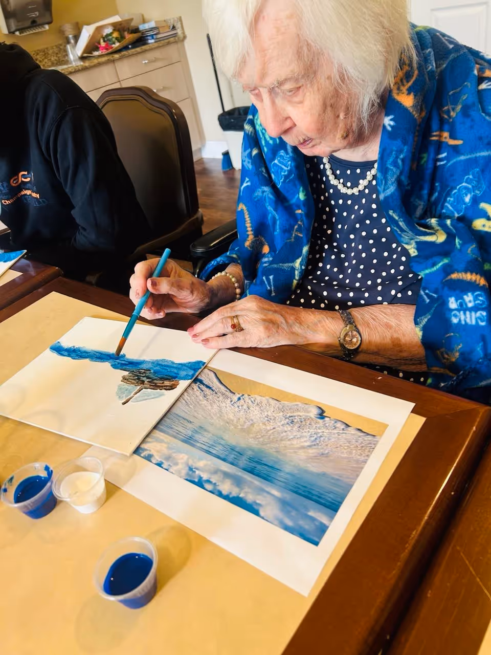 An elderly woman wearing a blue patterned shawl and a polka dot shirt is sitting at a table painting a picture of a seascape. She is using a paintbrush and has small containers of blue and white paint in front of her. There is a printed photo of a wave on the table that she appears to be using as a reference.
