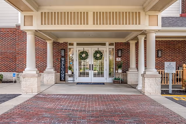Entrance of a senior living facility with double glass doors decorated with green wreaths, flanked by white columns and brick walls. A vertical welcome sign is placed on the left side of the doors, and a sign indicating a private residential community is on the right. The ground is paved with red bricks and concrete.