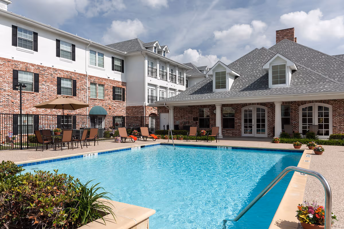 Outdoor swimming pool area at Twin Rivers Senior Living with lounge chairs, tables with umbrellas, and a brick building with white trim in the background under a partly cloudy sky.