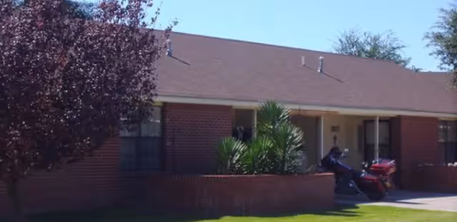 Single-story brick building with a brown shingled roof, a small tree and green shrubs in front, and a person sitting on a red mobility scooter near the entrance.