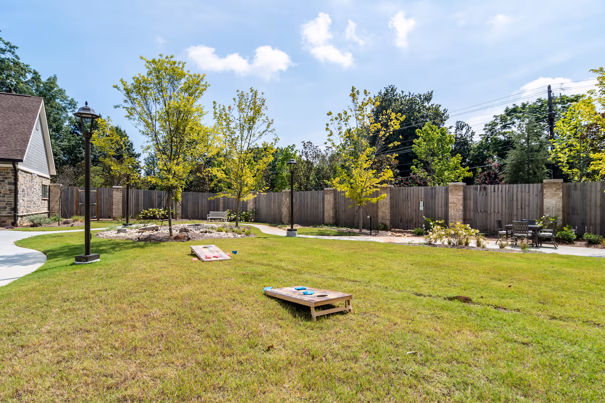 A sunny enclosed outdoor courtyard with grass, cornhole boards, young trees, benches, and a wooden fence.