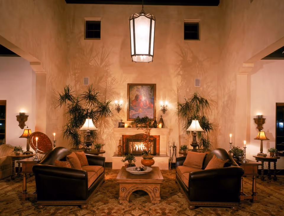 A warmly lit living room with two dark leather sofas facing each other across a wooden coffee table. The room features a fireplace with a fire burning, flanked by two tall plants and table lamps on either side. Above the fireplace is a framed painting, and the walls have sconces with candles. The ceiling has a large hanging lantern light fixture.