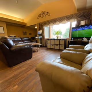 A cozy living room with leather sofas and armchairs arranged around a coffee table. There is a large window with curtains letting in natural light, a TV mounted on a stand showing a sports game, and decorative wall art above the window. The floor is wooden, and there is a storage unit with cubbies beneath the window.
