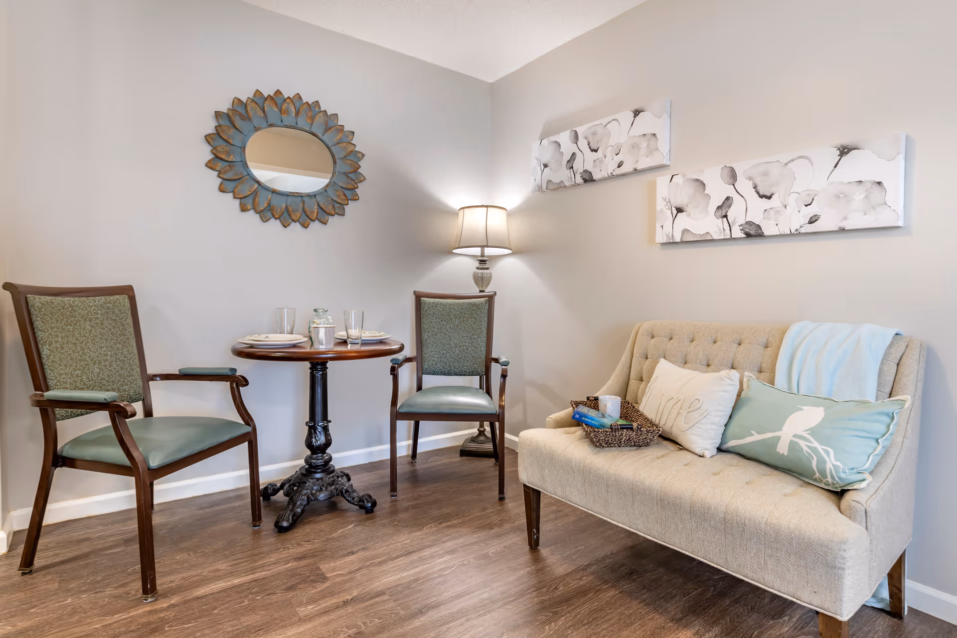 Cozy sitting area with two wooden chairs at a small round table, a tufted loveseat with pillows, wall art, and a floor lamp.