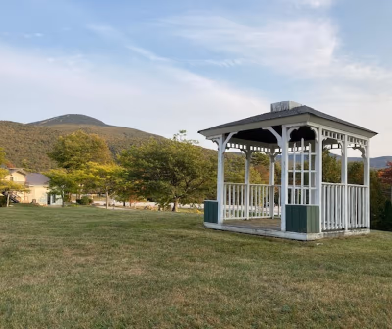 White wooden gazebo on a grassy lawn with trees, a nearby building, and hills in the background.