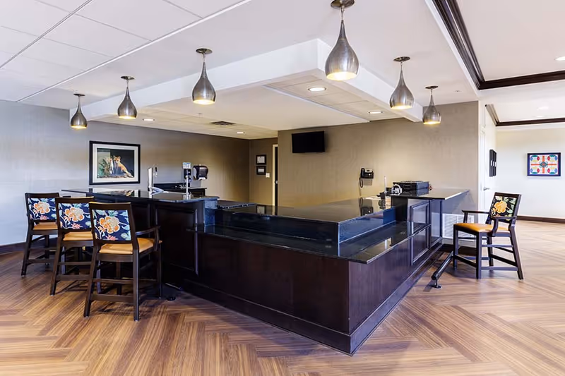 Interior view of a modern common area with a large dark wood and black granite counter surrounded by chairs with floral patterned cushions. The room has wood-patterned flooring, pendant lights hanging from the ceiling, a wall-mounted TV, and framed artwork on the walls.