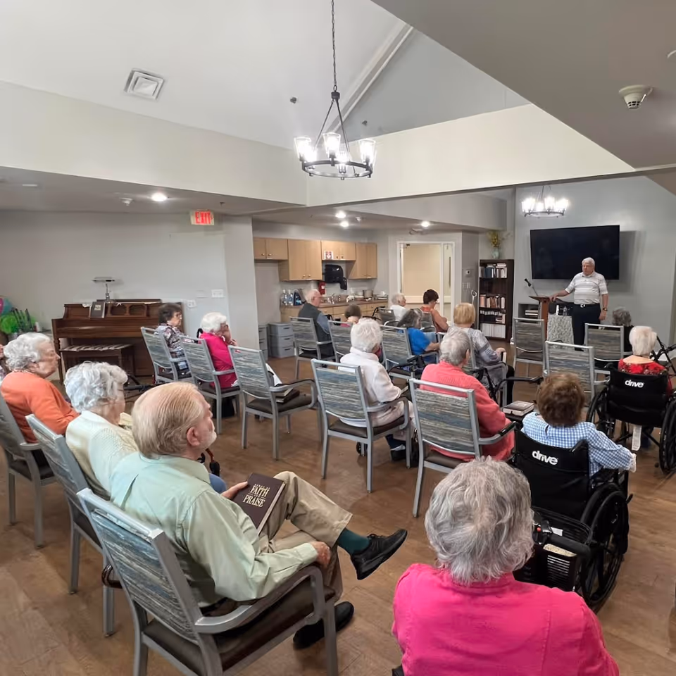 A group of elderly people seated in chairs and wheelchairs in a common room, attentively listening to a man standing near a TV screen at the front. The room has wooden flooring, a piano, and a kitchenette area in the background.