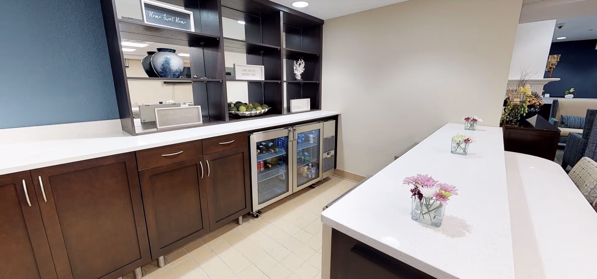 Long white countertop with small flower vases, dark wood cabinets and a glass-front beverage fridge in a senior living community serving/kitchenette area.