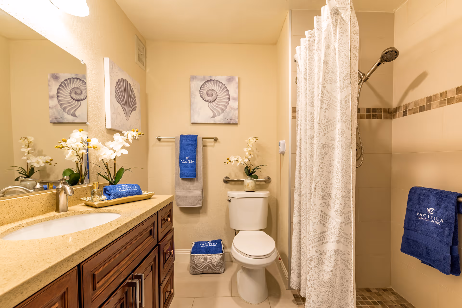 Bright tiled bathroom with a vanity and sink on the left, a toilet in the center, and a shower with a patterned curtain on the right, decorated with blue towels and shell artwork.