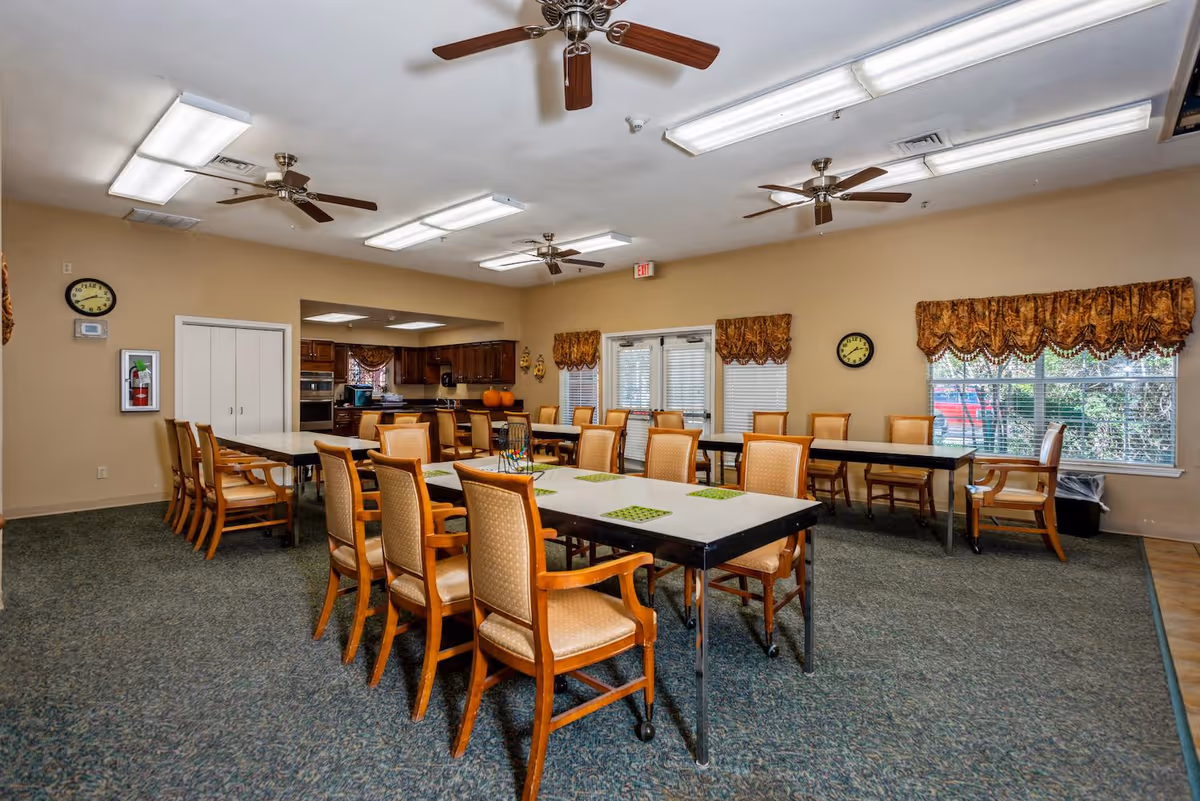 A spacious dining room with multiple tables and wooden chairs arranged neatly. The room has beige walls, carpeted flooring, ceiling fans, and fluorescent lighting. There are windows with patterned valances and a kitchen area visible in the background.
