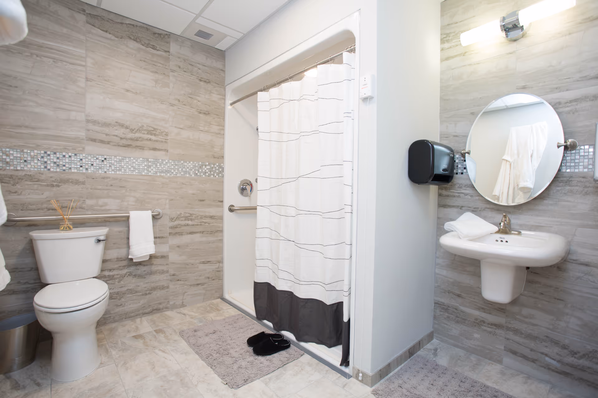 A clean and modern bathroom featuring a toilet with a towel hanging on a grab bar, a shower with a white curtain, a wall-mounted sink with a round mirror above it, a soap dispenser, and light gray tiled walls and floor. There are bath mats on the floor and a pair of black slippers near the shower.
