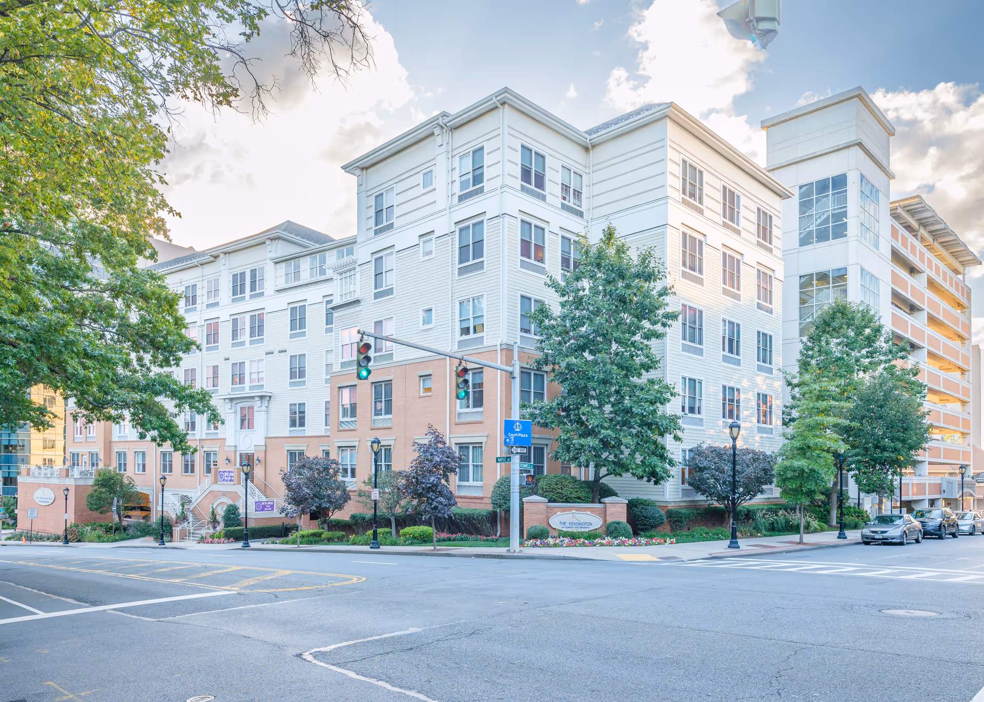 Exterior view of The Kensington White Plains, a multi-story residential building with a mix of brick and white siding. The building is situated at a street corner with traffic lights, trees, and parked cars visible. The sky is partly cloudy.