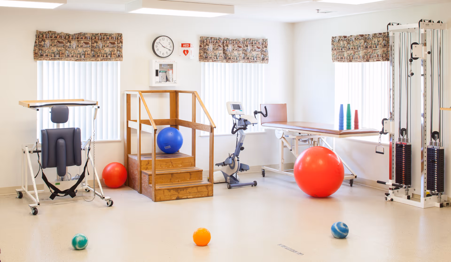 A bright physical therapy room with exercise equipment including a stationary bike, therapy table, pulley machine, wooden stairs with handrails, and various exercise balls scattered on the floor. The room has three windows with vertical blinds and patterned valances, a wall clock, and an AED mounted on the wall.