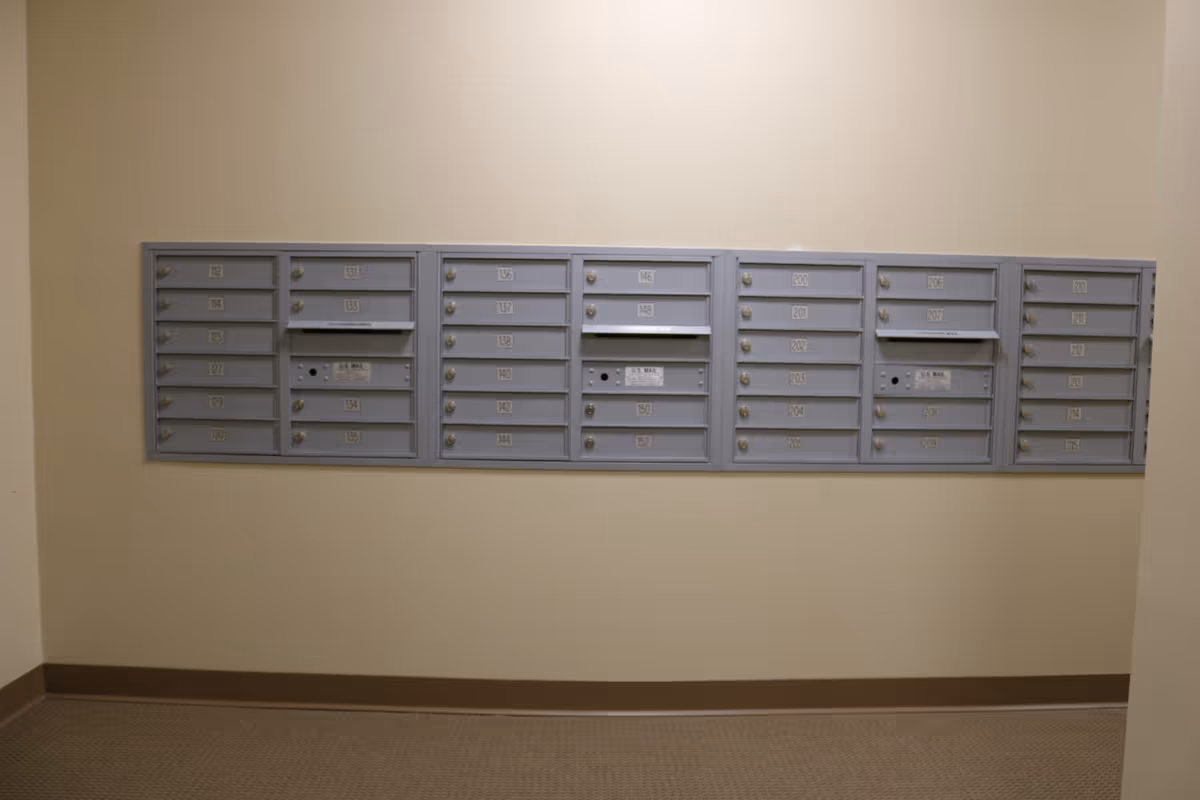 A row of gray metal mailboxes mounted on a beige wall in an indoor hallway, each mailbox labeled with numbers and some with mail slots.