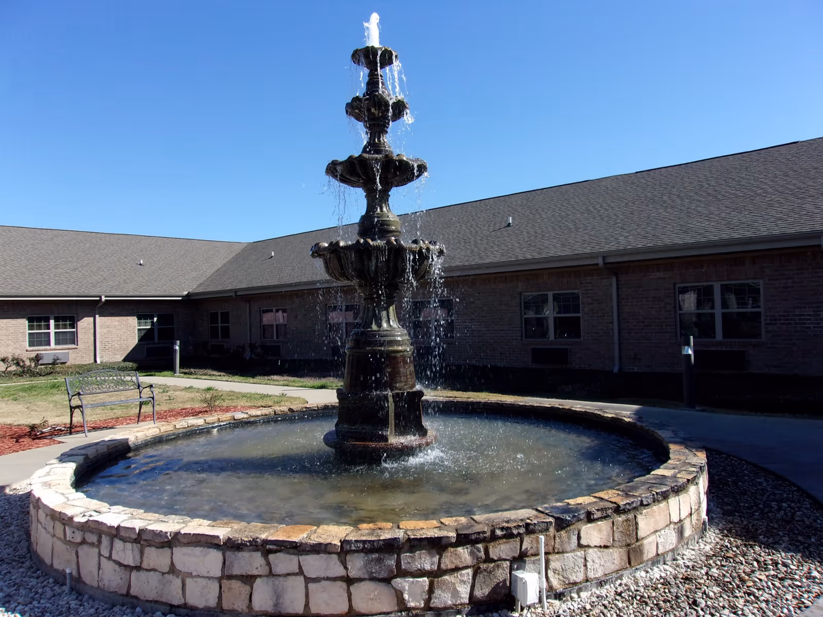 Outdoor courtyard area with a multi-tiered water fountain in the center, surrounded by a circular stone wall. There is a metal bench on the left side and a brick building with multiple windows in the background under a clear blue sky.